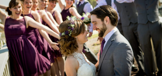 Photography of a young bride and groom looking each other in the eyes while the wedding party celebrates in the background.